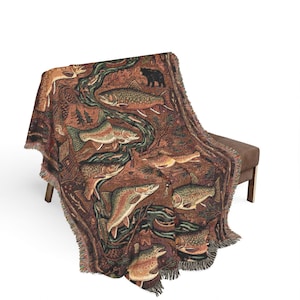May include: A woven throw blanket featuring a nature scene with fish, a bear, and a deer in shades of brown, green, and tan. The blanket has a fringed edge and is draped over a brown bench. The design includes a river and trees.