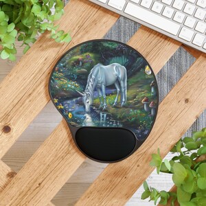 May include: A round, black ergonomic mousepad featuring a fantasy scene. A white unicorn drinks from a stream in a lush forest setting. The mousepad has a built-in wrist rest for added comfort.