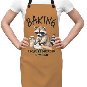 May include: Brown apron with black straps featuring a raccoon baking. The word "BAKING" is at the top, with "BECAUSE MURDER IS WRONG" below the raccoon. The raccoon is depicted with a jar, dough, and rolling pin.