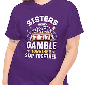 May include: A purple t-shirt with the text "Sisters That Gamble Together Stay Together". The graphic includes dice, poker chips, and a slot machine with triple sevens.