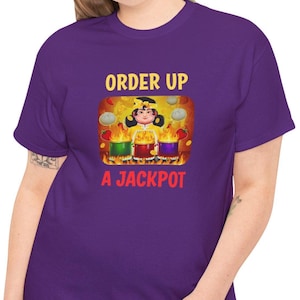 May include: Purple t-shirt with a graphic featuring a cartoon chef and the text "ORDER UP A JACKPOT." The chef is surrounded by pots with flames and food items. The shirt is made of a soft material.