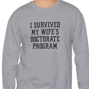 May include: A gray sweatshirt with the text "I SURVIVED MY WIFE'S DOCTORATE PROGRAM" in black block letters. The sweatshirt has a crew neck and long sleeves. The garment is made of a soft, comfortable material.