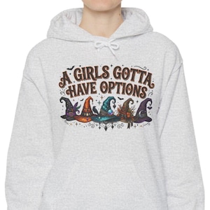 May include: A light gray hooded sweatshirt featuring a graphic print with the text A GIRLS GOTTA HAVE OPTIONS above five colorful, detailed witch hats in shades of purple, orange, teal, and black, accented with small stars, bats, and swirls.