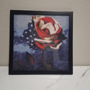 Puede incluir: Pintura de diamantes enmarcada de una rosa roja y blanca sobre un fondo azul. La obra de arte está compuesta por pequeñas gemas brillantes, creando un efecto de mosaico. El marco es negro.