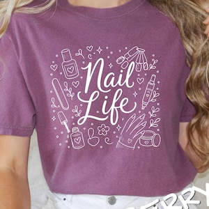 May include: A berry-colored short-sleeve t-shirt featuring a white graphic print with the text "Nail Life" surrounded by illustrations of nail polish bottles, a nail file, a cuticle oil pen, a hand, and small heart and flower accents.