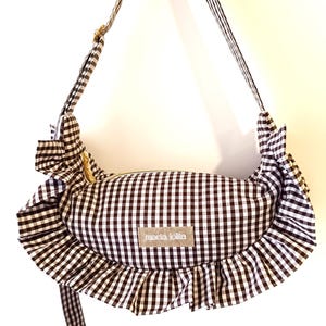 May include: A black and white gingham pattern crossbody bag with a ruffled edge. The bag has a gold zipper and a matching adjustable strap. The label "maria lolita" is visible on the front.