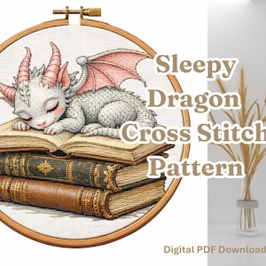 Peut inclure: Motif de point de croix d'un bébé dragon endormi aux détails roses, posé sur une pile de livres. Le motif est dans un cercle à broder en bois. Le texte indique "Sleepy Dragon Cross Stitch Pattern" et "Digital PDF Download."