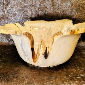 May include: A decorative wooden bowl with a unique, organic shape. The bowl features a light beige interior and a rim with a natural wood grain pattern. The exterior has a smooth, polished finish, showcasing the natural wood tones and textures.
