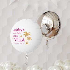 May include: A round white balloon featuring the pink text ashley's bombshells hit the VILLA Miami 2026 with gold palm tree and heart graphics, paired with a reflective silver metallic balloon, both attached to curled ribbons and floating in the air.