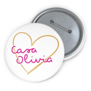 Könnte beinhalten: Weißer Ansteckbutton mit goldener Herzumrandung und dem pinkfarbenen Schriftzug "Casa Olivia". Der Button hat eine Metallnadel auf der Rückseite. Ein weiterer Button ist teilweise sichtbar.