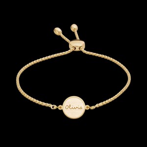 Puede incluir: Una pulsera ajustable de tono dorado con una cadena fina de eslabones cuadrados y un dije circular central. El disco tiene grabado el nombre Olivia en letra cursiva. La pulsera incluye un mecanismo de cuenta deslizante para ajustar.