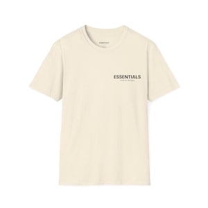 May include: Cream-coloured t-shirt with the word "ESSENTIALS" printed in black on the front. The shirt has a crew neck and short sleeves. The text "FEAR OF GOD" is printed below the word "ESSENTIALS".