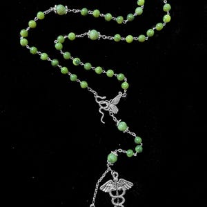 Op de afbeelding: Een rozenkransketting met groene kralen en zilverkleurige metalen accenten. De ketting heeft een vlinderbedel, een medisch symbool en een kleine ovale hanger. De kralen zijn lichtgroen.