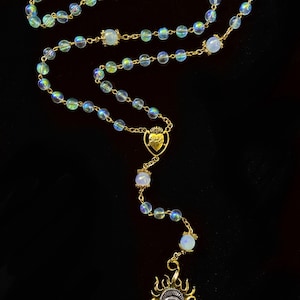 Può includere: Un rosario color oro con perline di vetro iridescenti e una catena dorata. Il rosario presenta un ciondolo a forma di cuore con una corona e un ciondolo a forma di sole con un disegno a occhio. Le perline sono un mix di colori chiari e iridescenti.