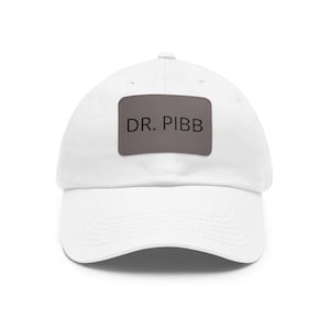 May include: White baseball cap with a curved brim. The cap features a rectangular patch with the text "DR. PIBB" in black letters. The hat is made of a soft material and is suitable for casual wear.
