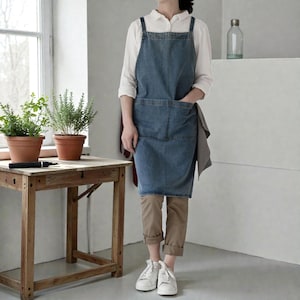 May include: A denim apron with a large front pocket and cross-back straps. The apron is a medium wash blue color. The person is wearing a white shirt and khaki pants. The apron is designed for kitchen or garden use.