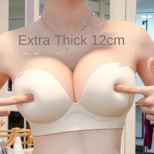 May include: A beige strapless bra with a deep V-neck design. The text "Extra Thick 12cm" is displayed above the bra. The bra is being pointed at by two fingers.