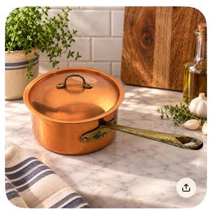May include: A round copper saucepan with a matching lid and a long, riveted brass handle. The pot features a polished metallic finish and a small, circular maker's mark on the handle attachment. The lid has a curved brass handle on top.