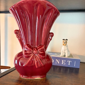 May include: A glossy, deep red ceramic vase featuring a flared, fan-shaped top and a decorative molded bow detail on the front. The vase has a rounded base and vertical ribbed textures extending from the center bow up to the wide, scalloped rim.