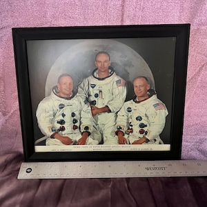 May include: A framed photograph featuring the prime crew of the Apollo 11 mission: Neil Armstrong, Michael Collins, and Edwin Aldrin Jr. The astronauts are in white spacesuits, standing in front of a large moon. The frame is black.