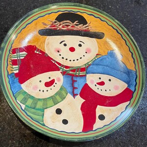 May include: A round ceramic plate featuring a painted illustration of three snowmen. The central snowman wears a black hat, while the two smaller snowmen wear red and blue knit caps. They are adorned with colorful scarves against a yellow background.