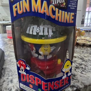 May include: A blue cardboard box containing an M&M's Fun Machine candy dispenser. The packaging features the text Fun Machine and Dispenser, with images of red and yellow M&M characters. It includes a 9.40 oz bag of milk chocolate candies.