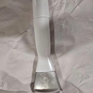 May include: A handheld kitchen tool featuring a smooth, cylindrical white handle that tapers toward a flared base. Attached to the bottom is a wide, metallic, silver-colored blade with a curved edge, designed for cutting or scraping tasks.