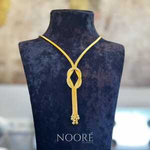 Puede incluir: Un collar dorado con un dise&ntilde;o de nudo y una borla de peque&ntilde;as cuentas. El collar se exhibe sobre un busto de terciopelo negro. La marca "NOOR&Eacute; ISTANBUL" est&aacute; impresa en la parte inferior.