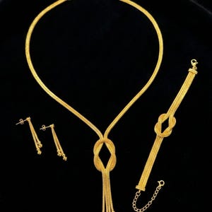 Puede incluir: Un conjunto de joyas de oro que incluye un collar, una pulsera y pendientes. El collar y la pulsera tienen un dise&ntilde;o anudado, y los pendientes son colgantes. El conjunto se muestra sobre un fondo negro. La marca "NOORE ISTANBUL" es visible.