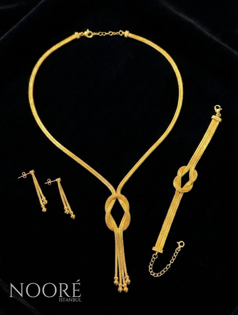 Puede incluir: Un conjunto de joyas de oro que incluye un collar, una pulsera y pendientes. El collar y la pulsera tienen un dise&ntilde;o anudado, y los pendientes son colgantes. El conjunto se muestra sobre un fondo negro. La marca "NOORE ISTANBUL" es visible.