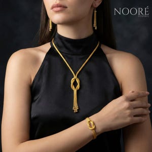 Puede incluir: Un conjunto de joyas de color dorado que incluye un collar, una pulsera y pendientes. El collar tiene un dise&ntilde;o de nudo con una cadena colgante. La pulsera y los pendientes tambi&eacute;n tienen un dise&ntilde;o de nudo. La marca "NOOR&Eacute; ISTANBUL" es visible.