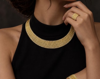 May include: A gold jewelry set featuring a wide, textured mesh necklace, a matching wide mesh bracelet, a wide mesh ring, and rectangular drop earrings with a fine beaded texture. The set is displayed against a black background with the text NOORÉ ISTANBUL.