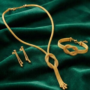 Puede incluir: Un conjunto de joyas de oro que incluye un collar, una pulsera y pendientes, exhibido sobre una superficie de terciopelo verde. El collar presenta un dise&ntilde;o de nudo y una cadena colgante. La pulsera tambi&eacute;n tiene un dise&ntilde;o de nudo. Los pendientes tienen un dise&ntilde;o de bola colgante. El logotipo de NOOR&Eacute; ISTANBUL es visible.