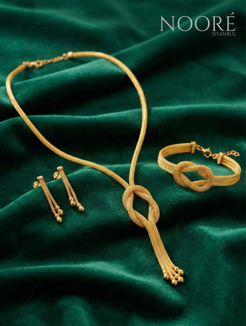 Puede incluir: Un conjunto de joyas de oro que incluye un collar, una pulsera y pendientes, exhibido sobre una superficie de terciopelo verde. El collar presenta un dise&ntilde;o de nudo y una cadena colgante. La pulsera tambi&eacute;n tiene un dise&ntilde;o de nudo. Los pendientes tienen un dise&ntilde;o de bola colgante. El logotipo de NOOR&Eacute; ISTANBUL es visible.