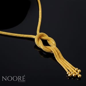 Puede incluir: Un collar dorado con un dise&ntilde;o de nudo y hebras colgantes que terminan en peque&ntilde;as esferas. El collar se muestra sobre un fondo negro. La marca "NOOR&Eacute; ISTANBUL" es visible en la parte inferior.