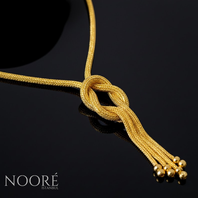 Puede incluir: Un collar dorado con un dise&ntilde;o de nudo y hebras colgantes que terminan en peque&ntilde;as esferas. El collar se muestra sobre un fondo negro. La marca "NOOR&Eacute; ISTANBUL" es visible en la parte inferior.