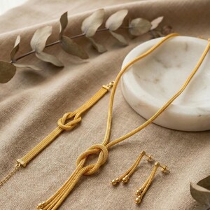 Puede incluir: Un conjunto de joyas de color dorado que incluye un collar, una pulsera y pendientes. El collar y la pulsera tienen un dise&ntilde;o de nudo, y los pendientes tienen cuentas colgantes. Las joyas se exhiben sobre una tela de tono neutro.