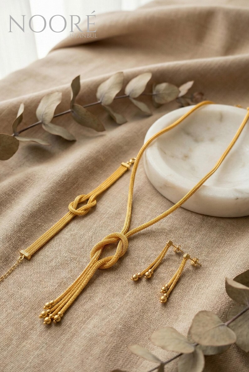 Puede incluir: Un conjunto de joyas de color dorado que incluye un collar, una pulsera y pendientes. El collar y la pulsera tienen un dise&ntilde;o de nudo, y los pendientes tienen cuentas colgantes. Las joyas se exhiben sobre una tela de tono neutro.