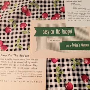May include: Three vintage recipe cards from Today's Woman Magazine featuring the text Easy on the Budget and Glazed Meat Loaf. The cards are displayed on a black and white gingham fabric background with a repeating pattern of red cherries and green leaves.