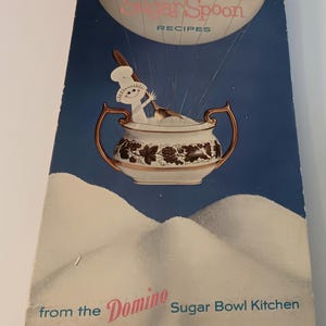 May include: Vintage spiral-bound cookbook titled "Sugar Spoon Recipes" from the Domino Sugar Bowl Kitchen. The cover features a whimsical illustration of a sugar bowl with a chef stirring sugar. The background is blue with white snow-like shapes.