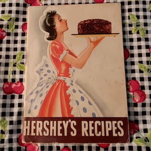 May include: A vintage-style cookbook titled HERSHEY'S RECIPES featuring an illustration of a person in a red dress and a white apron with blue polka dots holding a tray with a large chocolate cake. The cover has a cream background.
