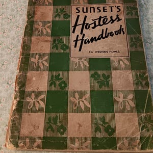 May include: A vintage book titled Sunset's Hostess Handbook For Western Homes. The cover features a green and beige checkered pattern with small floral motifs. The book shows signs of wear along the edges and spine, with a rectangular title box.