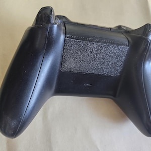 Könnte beinhalten: Ein schwarzer Xbox-Controller, von hinten betrachtet, mit Batteriefach und Griffen. Der Controller hat eine strukturierte Oberfläche auf der Batterieabdeckung und glatte, geschwungene Griffe. Das Design ist ergonomisch und elegant.