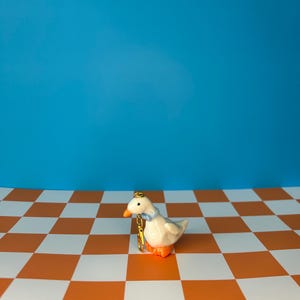 May include: A ceramic goose figurine with a gold crown and a blue bow tie. The goose has orange feet and a gold chain. It is set against a blue background and a checkered orange and white surface.