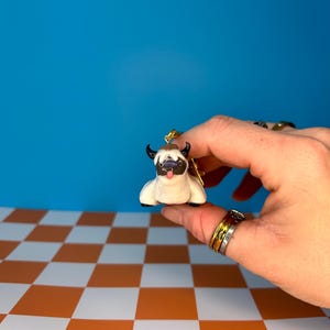 May include: A small, white and black figurine of a bull with black horns and a pink tongue. The bull figurine is held in a hand against a blue background and an orange and white checkered surface.