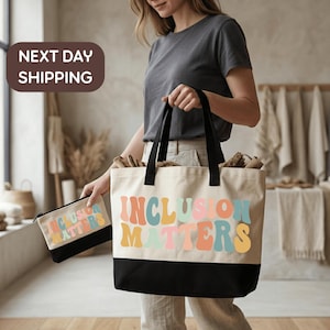May include: A cream-colored canvas tote bag and a matching zippered pouch, both featuring a black bottom panel and the text "INCLUSION MATTERS" printed in a wavy, multi-colored font with shades of pink, orange, green, and blue.