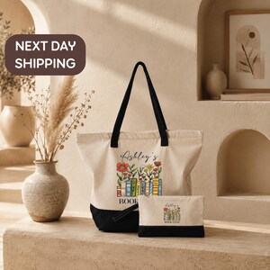 a tote bag and a small book bag with the text "Ashley's BOOK CLUB" printed on them, placed on a table with a vase of dried flowers and a framed picture in the background.