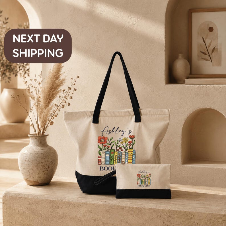 a tote bag and a small book bag with the text "Ashley's BOOK CLUB" printed on them, placed on a table with a vase of dried flowers and a framed picture in the background.