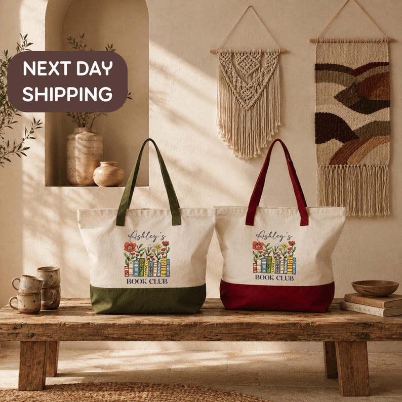 two tote bags with the text "Ashley's BOOK CLUB" printed on them, placed on a wooden bench. The bags are green and red in color. The background includes a wall with a hanging tapestry and a shelf with various decorative items. There is also a text overlay that reads "NEXT DAY SHIPPING".