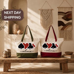 May include: Two off-white canvas tote bags featuring a graphic of four playing card suits in black and red, with the name Olivia written in black script below. One bag has an olive green base, and the other has a deep red base with matching handles.